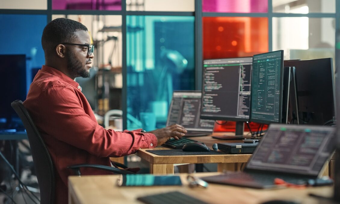 african american man writing lines of code on desktop computer with multiple monitors and laptop in creative office. male data scientist working on innovative online service for start up company.